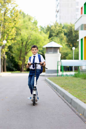 a handsome stylish and young schoolboy in a white shirt, blue tie and a backpack with a scooter on a streetの写真素材