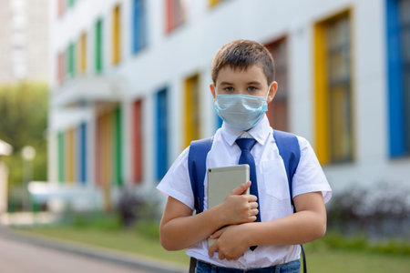 a schoolboy in a white shirt, blue tie and a backpack stands in a medical mask with a tablet
