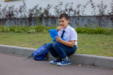 schoolboy in a white shirt with a blue tie, holds a blue lunch box and eats a piece of appleの写真素材
