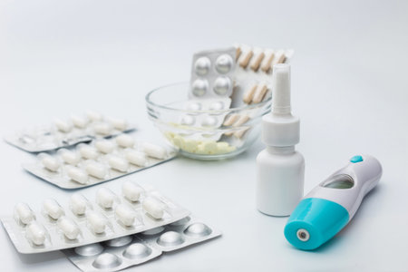 Silvery blisters and tablets, a bottle of nasal spray and an electronic thermometer lie on a white...