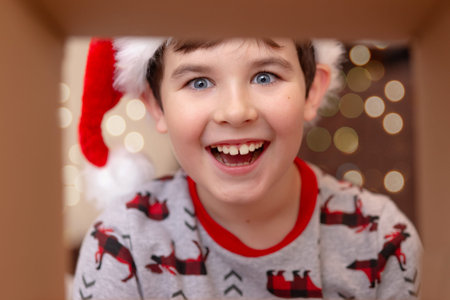 A surprised, happy, brunette boy in a Santa Claus hat takes a present out of the box