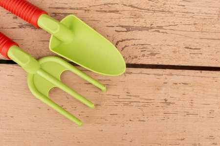 Light green garden hand tools, a shovel and a rake with orange handles, lie on painted boards.