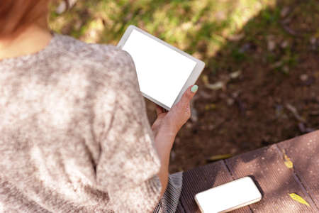 Girl sitting in a knitted jumper in the park holding a digital tablet and smartphone with a white screenの写真素材