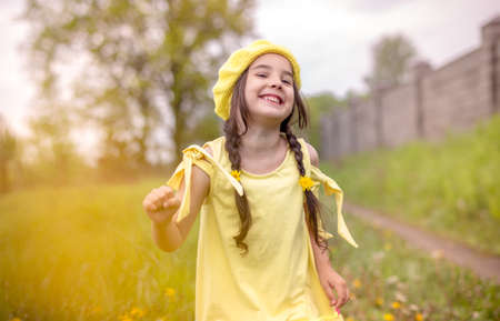 Cheerful and happy charming little girl in a yellow knitted beret and a yellow dress, dancing in nature in the springの写真素材