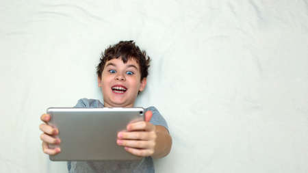 A funny brunette teenage boy lies on his back hold gray digital tabletの写真素材