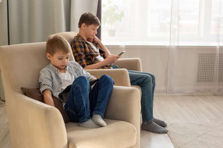 Two boys, a child and a teenager, sit in beige armchairs, in a room, during the day, play their gadgetsの写真素材