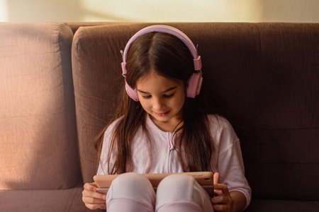 A beautiful little girl in pink headphones sits on the couch, looks into a digital tablet.