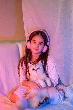 A beautiful little girl in pink headphones, with loose dark hair, sits with a white adorable catの写真素材