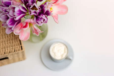 A bouquet of open pink and purple tulips, a wicker basket, a white cup with coffee on a white table.の写真素材