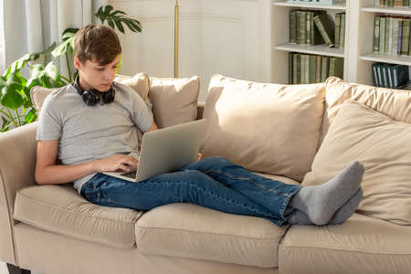 A teenager lying on a beige sofa, in a sunny room, wearing black headphones around his neck, looks into a laptop.の写真素材