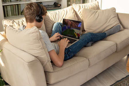 A teenager lying on a sofa in a room, wearing black headphones on his head, looks into a gray laptop, on his knees.の写真素材