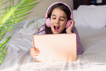 Happy little girl in pink headphones lying on a white bed, holding a pink digital tablet
