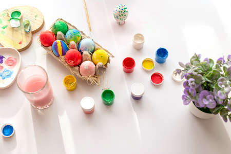 On a white table, in daylight, there are brushes in a glass of water, multi-colored eggs in a tray, jars of paint.の写真素材