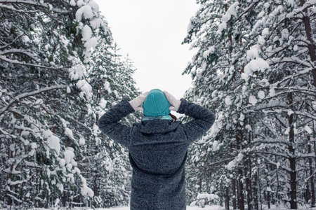 A girl in a gray coat and a light blue knitted hat stands with her hands to her head in a beautiful winter snowy forestの写真素材