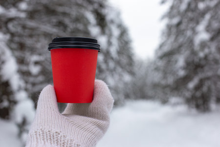 Red paper cup of coffee in hand in a white knitted mittens, against the backdrop of a beautiful winter forestの写真素材
