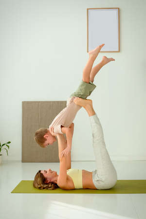 A woman, doing yoga with a child boy, lies on a sports mat, raising her legs up, doing exercises with support.の写真素材
