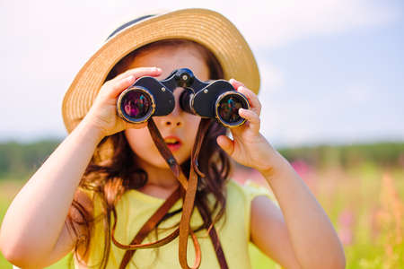 cute girl 4-5 years old, in a straw hat, stands in nature, against the blue sky, looks through binoculars to the sideの写真素材