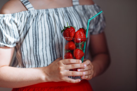 The hands of a girl in a striped blouse and a red skirt hold a glass with juicy strawberries.の写真素材