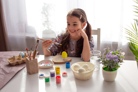 Charming little girl sits at a white table by the window, smiles, holds a brush, a painted egg stands in front of her.の写真素材