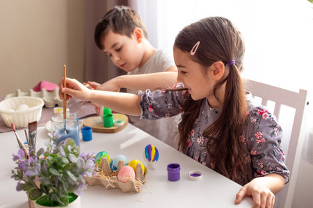 A cute little girl and boy sits at a white table near the window, paints eggsの写真素材