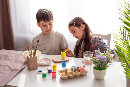 A brunette little girl and boy sits at a white table near the window, paints eggsの写真素材