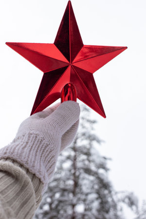 A hand in a white knitted mitten holds a red star over a snow pine tree in the forestの写真素材