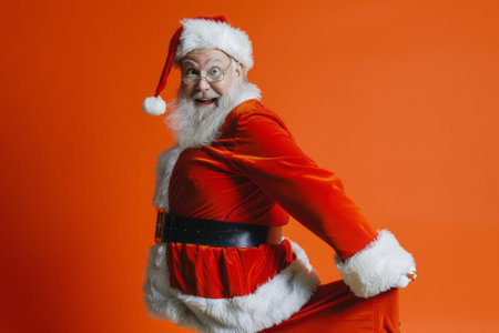 Cheerful santa in red suit with playful pose on festive orange background. Copy spaceの素材