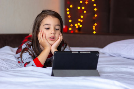 Little brunette girl in pajamas lies on the bed at home, looks into the tabletの写真素材