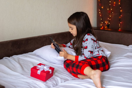 Little brunette girl in pajamas lies on the bed at home, looks into the digital tabletの写真素材