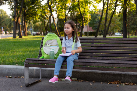 Young girl enjoying outdoor park with notepad and backpack on benchの写真素材