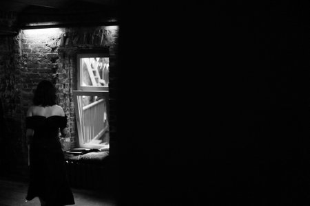 A girl in a black dress stands by the window in a dark room.