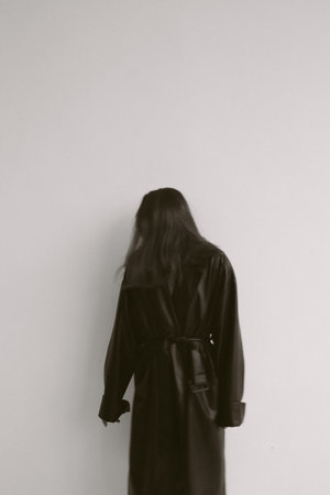Young woman in long black coat with hood on white wall background.の写真素材