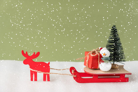 Toy reindeer and sleigh delivering Christmas present and tree on green background.