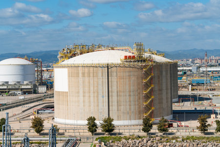 Massive round industrial storage tanks at a petrochemical plant. Visible pipelines, safety structures and distant city buildingsの写真素材