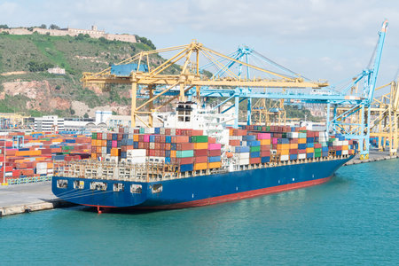 Container ship docked at busy port with cranes and hillside viewの写真素材