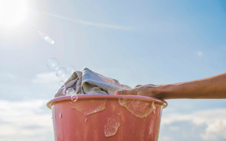 Female Hand washing clothes in the pink basin with clear Bubble soap against blue sky and sunlight.の素材
