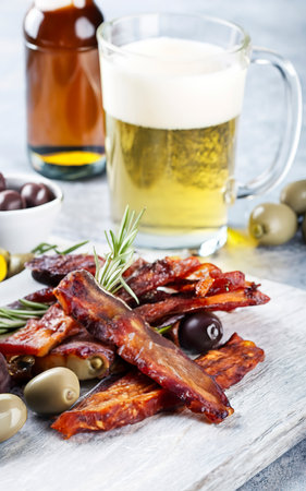 Jerky meat strips with spices, olives, rosemary and beer on wooden plate on a grey stone backgroundの素材