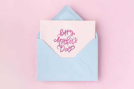 a pink card with 'happy mother's day' written in pink cursive script is partially pulled out of a light blue envelope, which is sitting on a solid light pink background