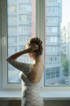beautiful girl with long brown hair in a white dress standing at the window from the back and corrects hairstyleの写真素材