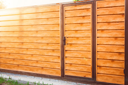 Fragment of a wooden brown modern fence with an entrance door In bright sunlight , closeup. Modern...