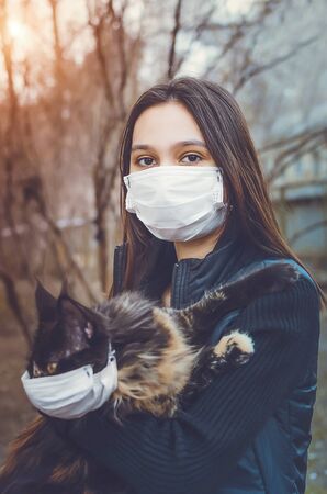 A girl in a medical mask on the street holds a cat in her arms. Cat in a protective mask. The concept of protection against coronavirus during an epidemic. Vertical tinted photo.の写真素材