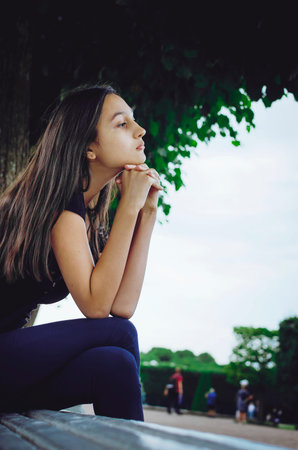 Pensive beautiful girl with long dark hair is sitting on a park bench.の写真素材