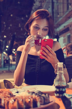 The girl drinks juice and reads the news in the smartphone while sitting in a cafe. Lifestyle, toned photography. Blurred background, soft focus.の写真素材