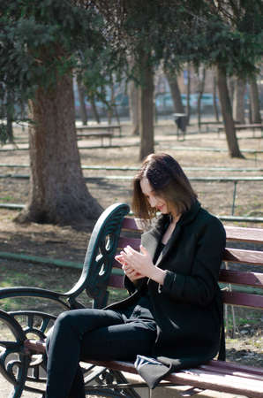 A beautiful happy girl uses a mobile phone while sitting on a park bench. vertical photo.の写真素材