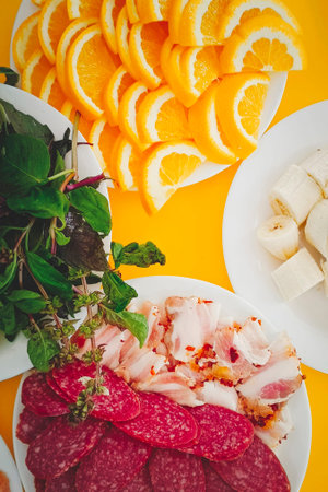 Sausage, bacon, basil greens, sliced orange lie on a plate on a served table. Vertical photo.