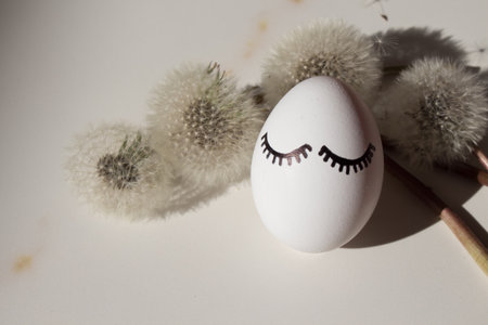 Stylish Easter egg with eyes and eyelashes on marble in dry dandelions with shadow. Fashion and Easter in harmony. Happy Easter card.の写真素材