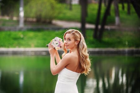 Young beautiful bride in a white dress with a bouquet standing near a pond waiting for the groomの写真素材