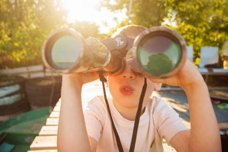 Portrait of little boy looking through binoculars on river bank. Cute kid with binoculars sitting on the pier on river. Focus on child.の写真素材