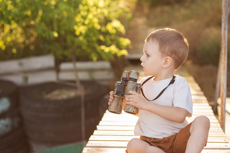Portrait of little boy looking through binoculars on river bank. Cute kid with binoculars sitting on the wooden pier on river.の写真素材