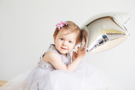 Portrait of a lovely little girl wearing elegant gray dress in front of a white background. Little princess. Little baby girl holding silver star-shaped balloon.の写真素材